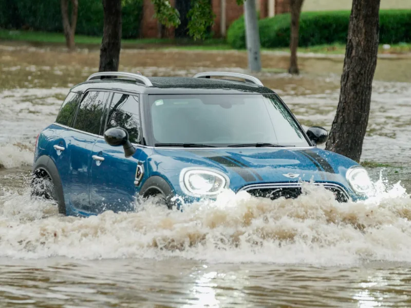 Ein blauer Kombi fährt durch eine überschwemmte Strasse. Hohe Wasserstände umgeben das Fahrzeug, während es durch die Wellen fährt. Bäume sind im Hintergrund zu sehen.