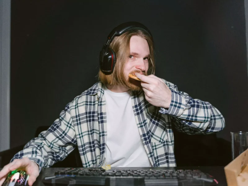 Ein junger Mann mit Kopfhörern sitzt vor einem Computer und isst ein Nahrungsmittel. Auf dem Tisch sind ein Gaming-Keyboard, eine Maus und eine Schüssel sichtbar. Der Hintergrund ist dunkel.