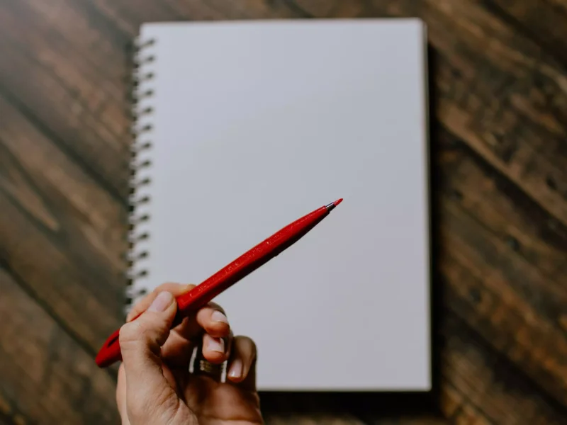 Eine Hand hält einen roten Stift über einem leeren Notizbuch auf einem hölzernen Tisch. Die Spiralbindung des Notizbuchs ist gut sichtbar.
