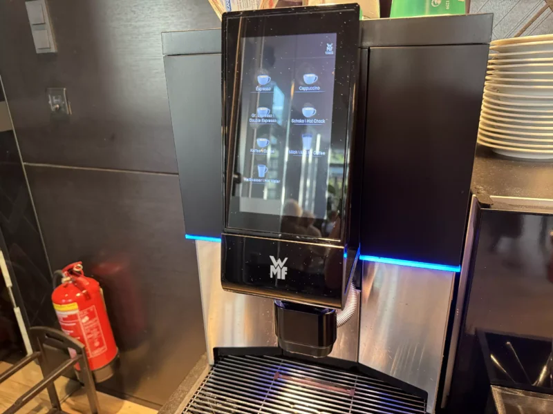 Ein moderner Kaffeeautomat mit Touchscreen, der verschiedene Getränkeoptionen wie Espresso, Cappuccino und heisse Schokolade anbietet. Im Hintergrund ist ein Feuerlöscher zu sehen.