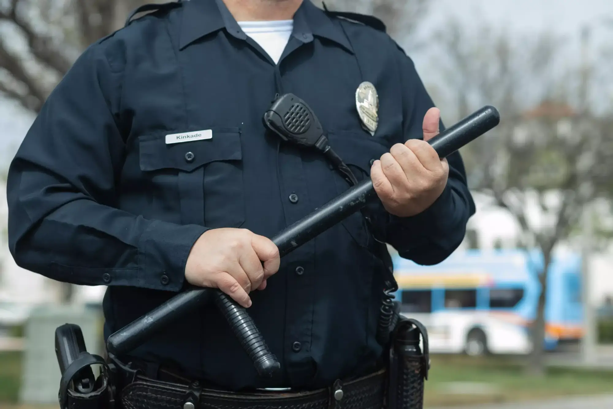Ein Polizist in Uniform hält einen Stock in der Hand. Im Hintergrund sind Bäume und ein Bus zu sehen. Die Szenerie vermittelt ein Gefühl von Sicherheit und Präsenz der Polizei.